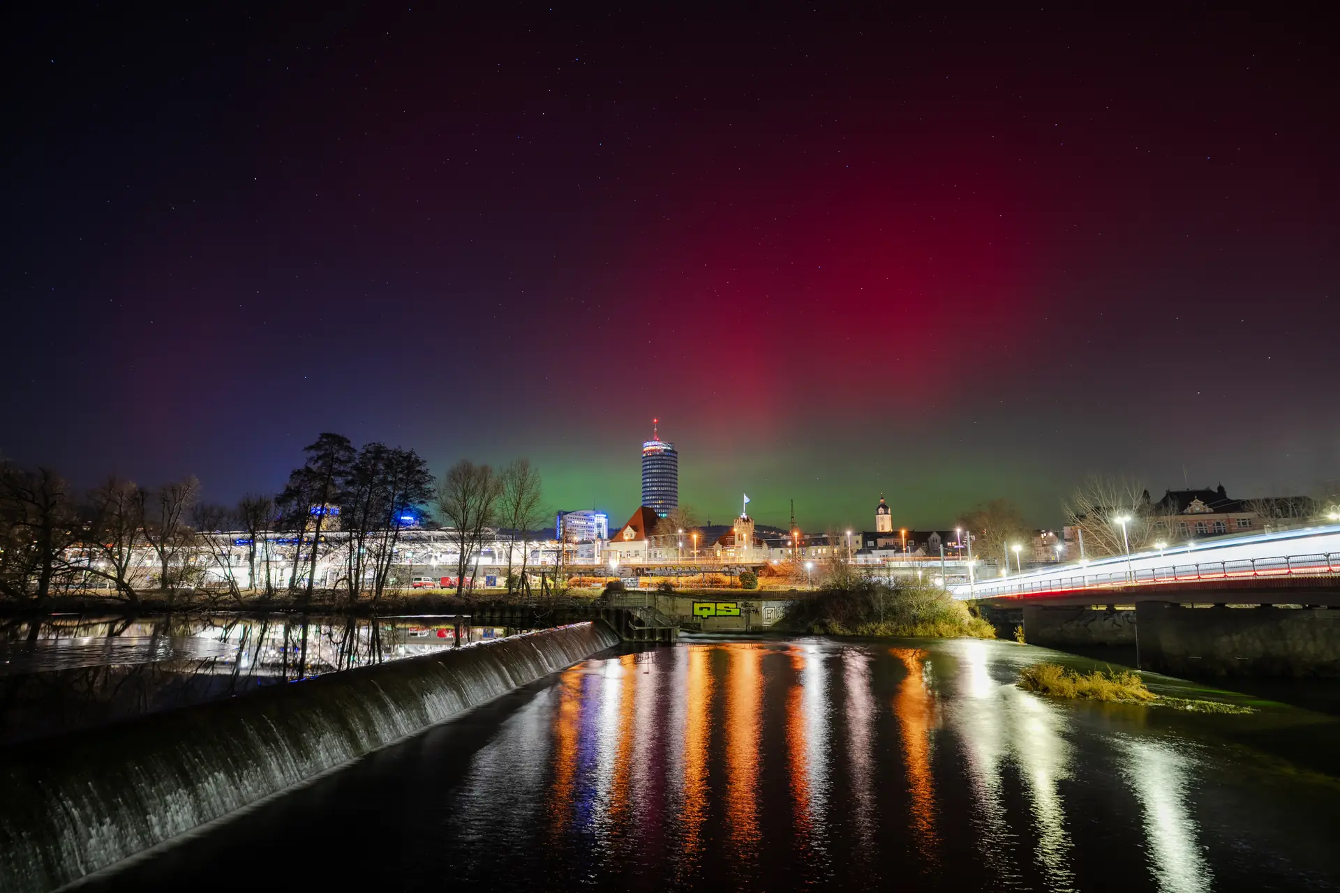 Rote und grüne Polarlichter über Jena, davor der JenTower und die Saale mit Wehr bei Nacht